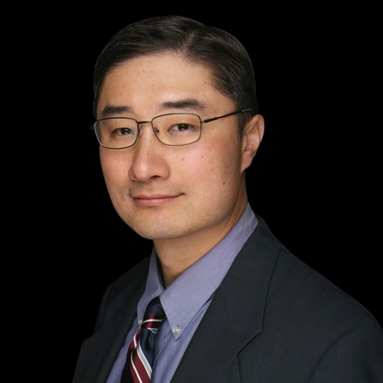man in glasses and blue shirt and printed tie and black suit coat in black background